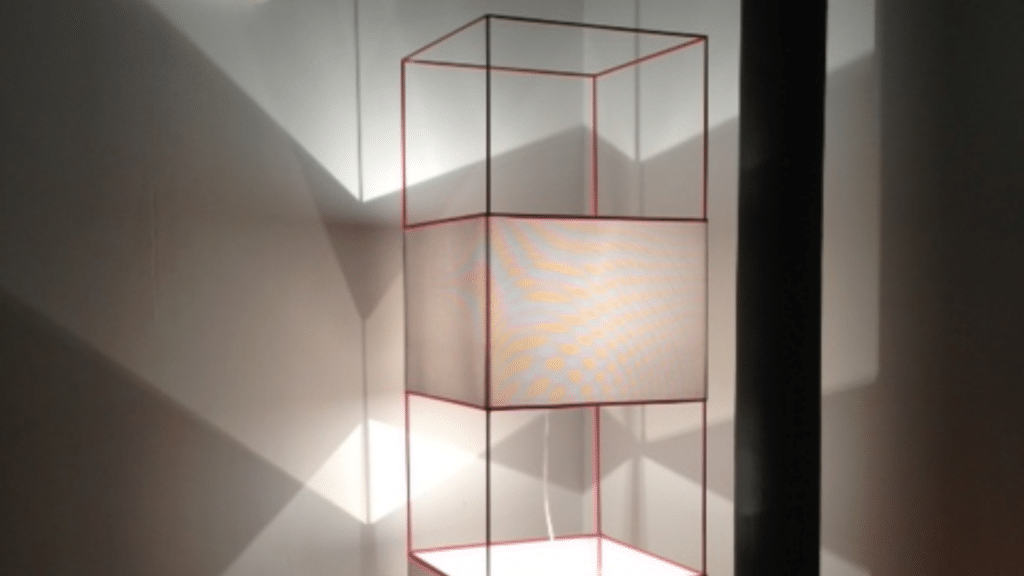Lampadaire à structure en métal rouge et abat-jour blanc, composé de cubes superposés, projetant un jeu d’ombres graphiques sur le mur